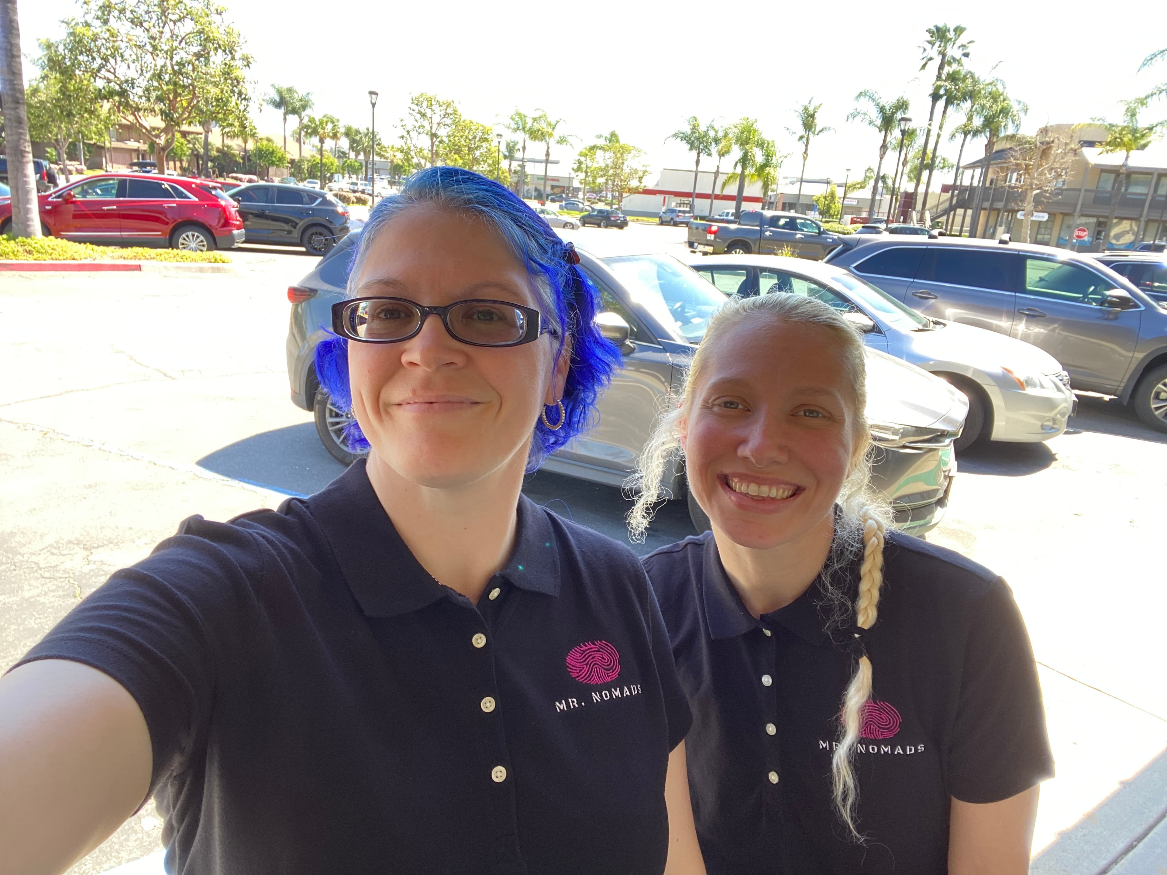 Upside-down selfie of two people wearing matching black MR. NOMADS polo shirts outdoors.
