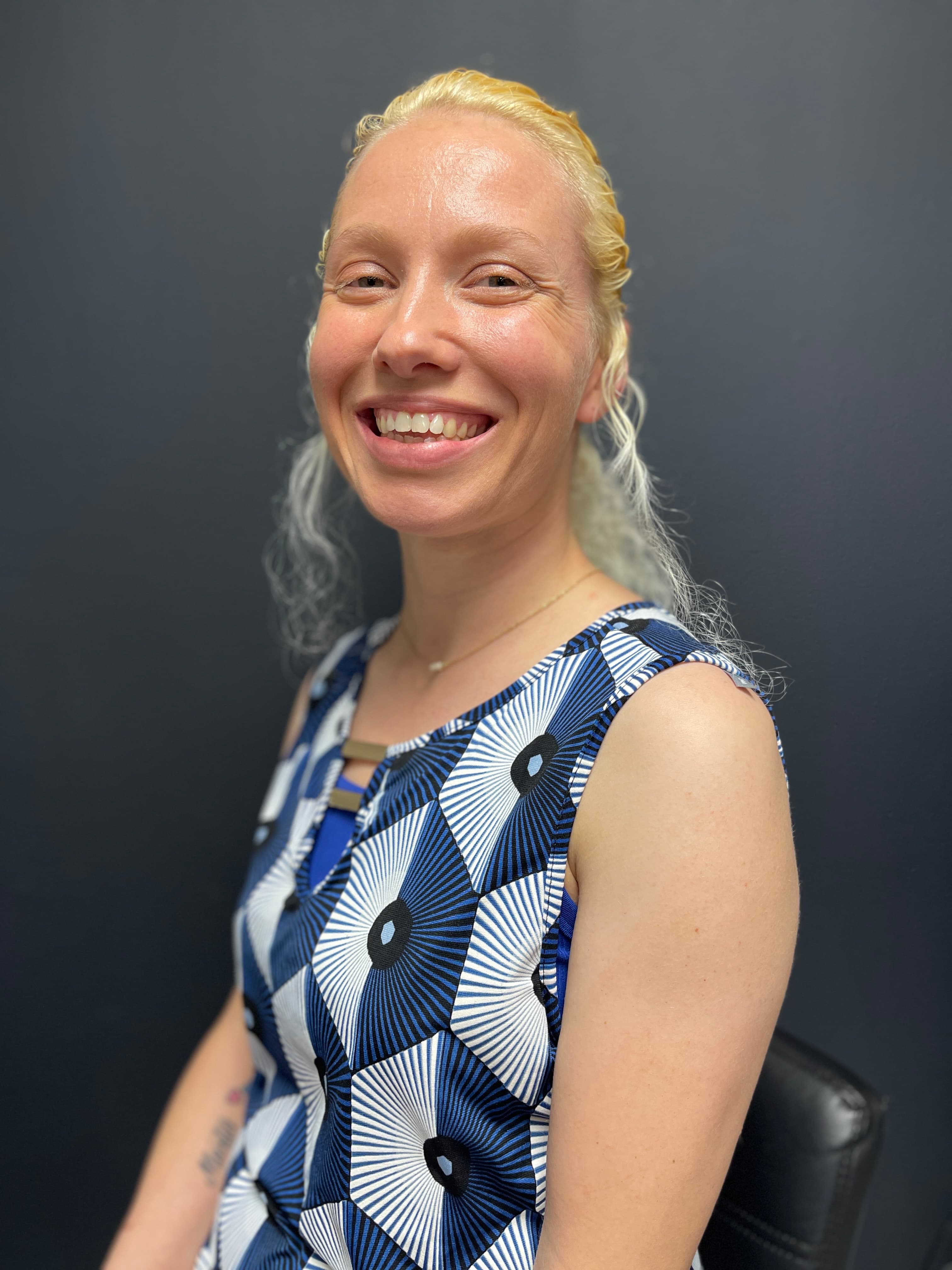 Smiling blonde woman wearing a blue and white patterned top against a dark background.