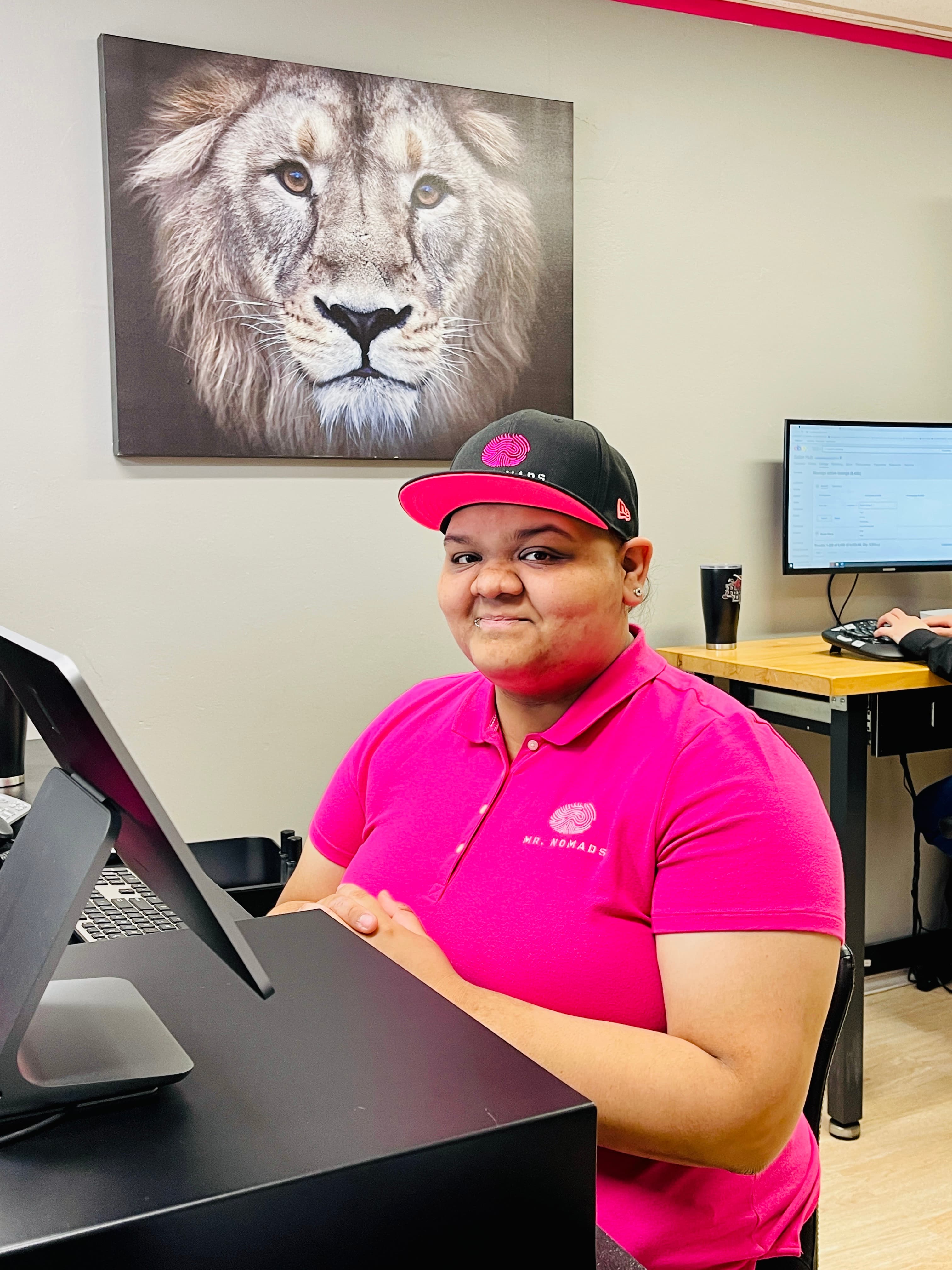 Smiling person in pink polo and cap sits at a desk with a lion portrait.