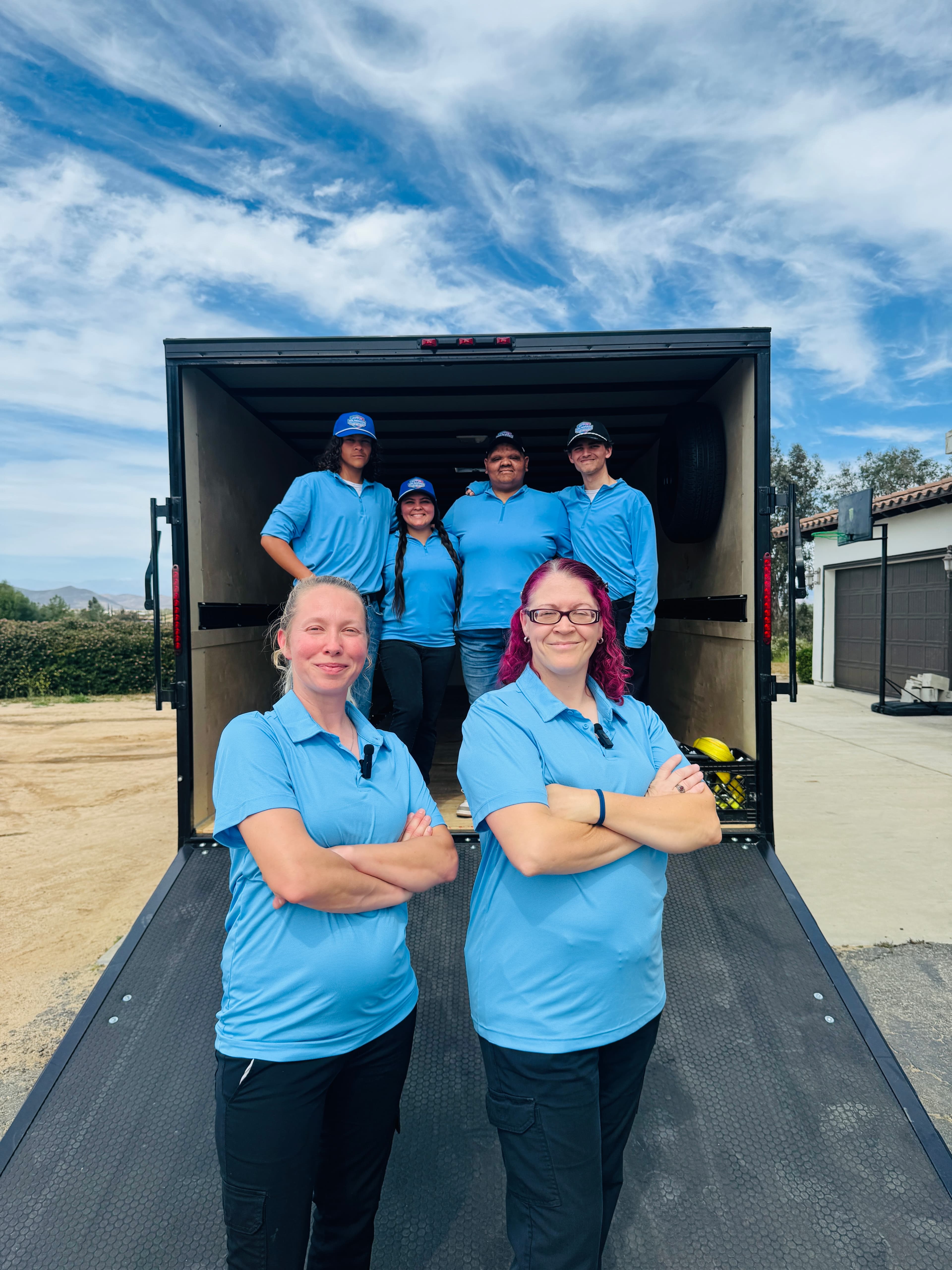 Six team members in blue uniforms posing in front of an open black cargo trailer.